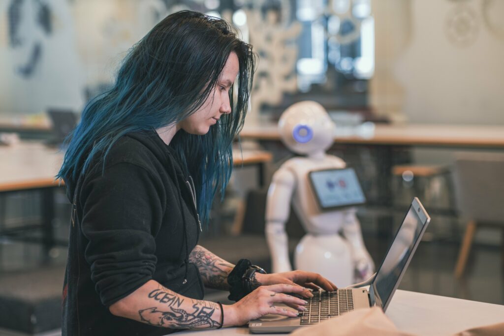 Pitkähiuksinen ihminen näppäilee kannettavaa tietokonetta. Hänellä on tatuoidut kädet. Taustalla seisoo Pepper-humanoidirobotti.