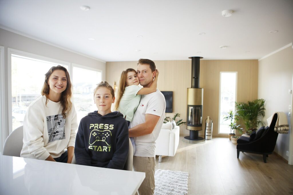 A boy standing by the table in the living room, his mother smiling next to him. Behind the boy, the father is holding the daughter of the family in his arms. A fireplace can be seen in the background.