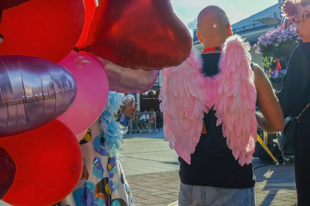 Outside in summer weather, red heart-shaped balloons in a bouquet and a man with his back to the camera. The man has pink angel wings on his back over a black t-shirt.