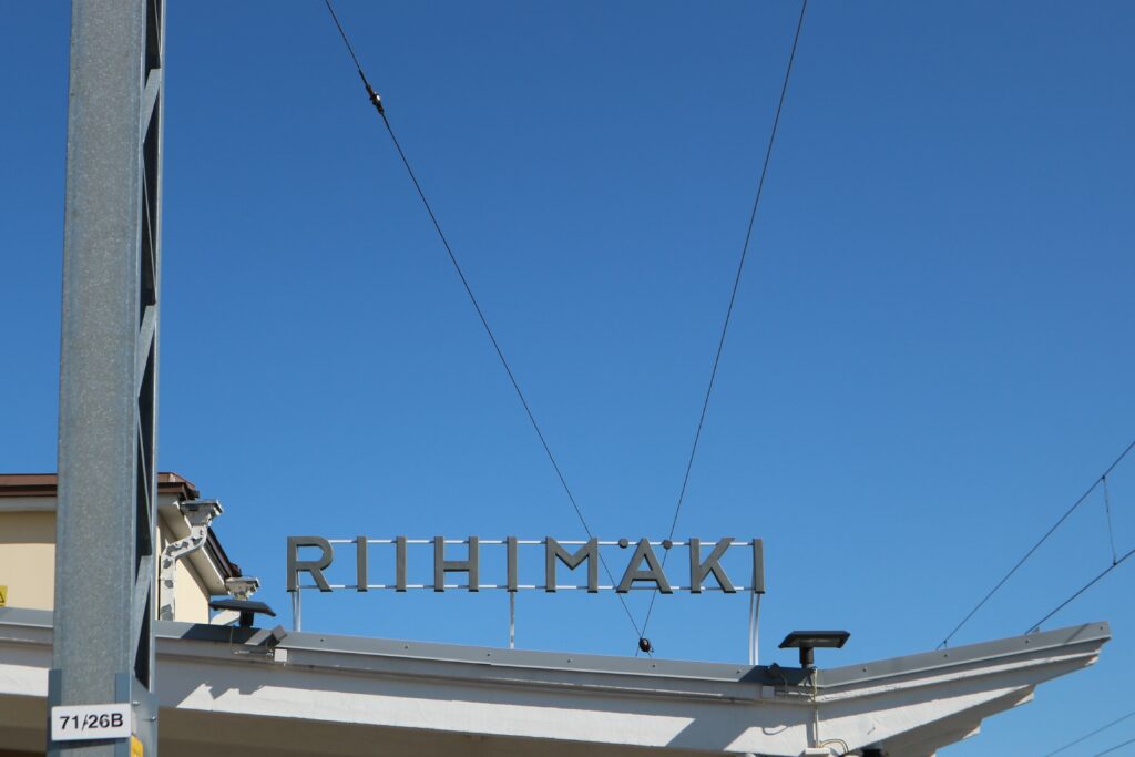 Rautatieaseman lipan päällä Riihimäki-kirjaimet vaaleansinistä taivasta vasten.