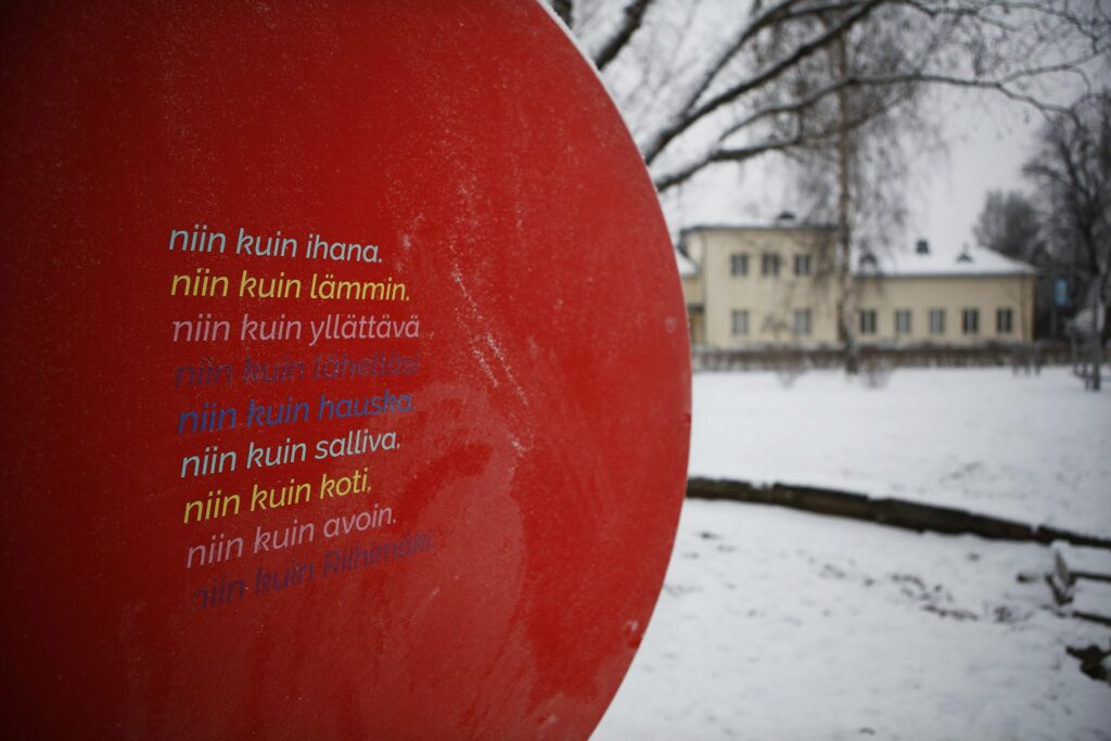 Talvisessa puistomaisemassa punainen suuri R-kirjainveistos, jonka kyljessä värikästä tekstiä.
