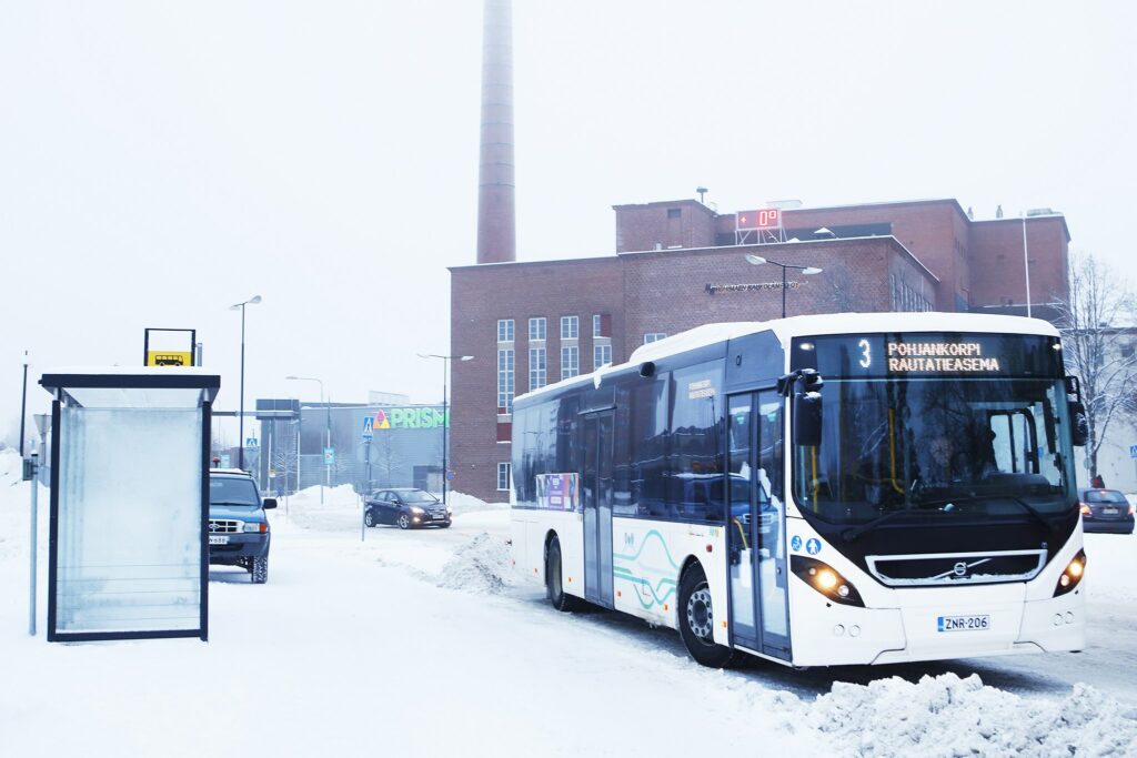 Bussi seisahtuneena pysäkille talvisessa maisemassa taustallaan vanha punatiilinen voimalarakennus.