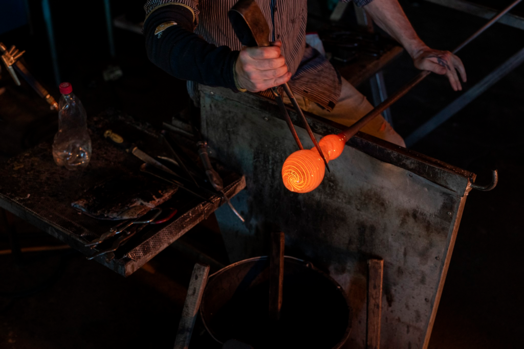 Hämärässä valaistuksessa lasinpuhaltaja muotoilemassa pajassaan pihdeillä kuumaa oranssina hehkuvaa lasia, joka on kiinni tangon päässä.