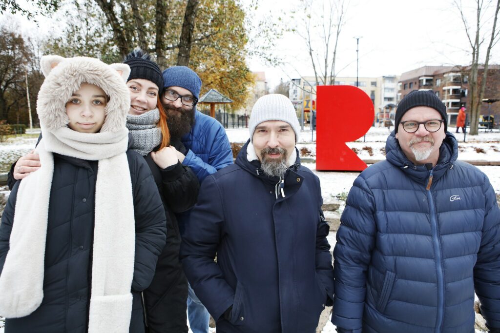 Two women and three men in their outerwear in a winter park with a red R-shaped sculpture in the background.