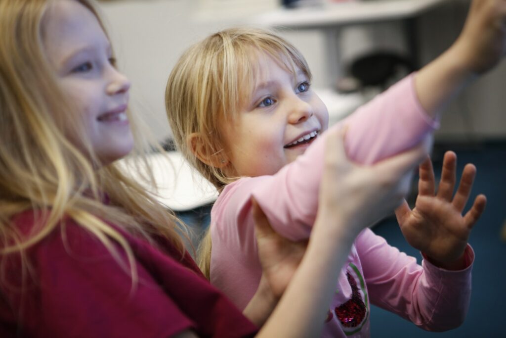 Two happy girls close-up in class, pointing and looking in common direction.