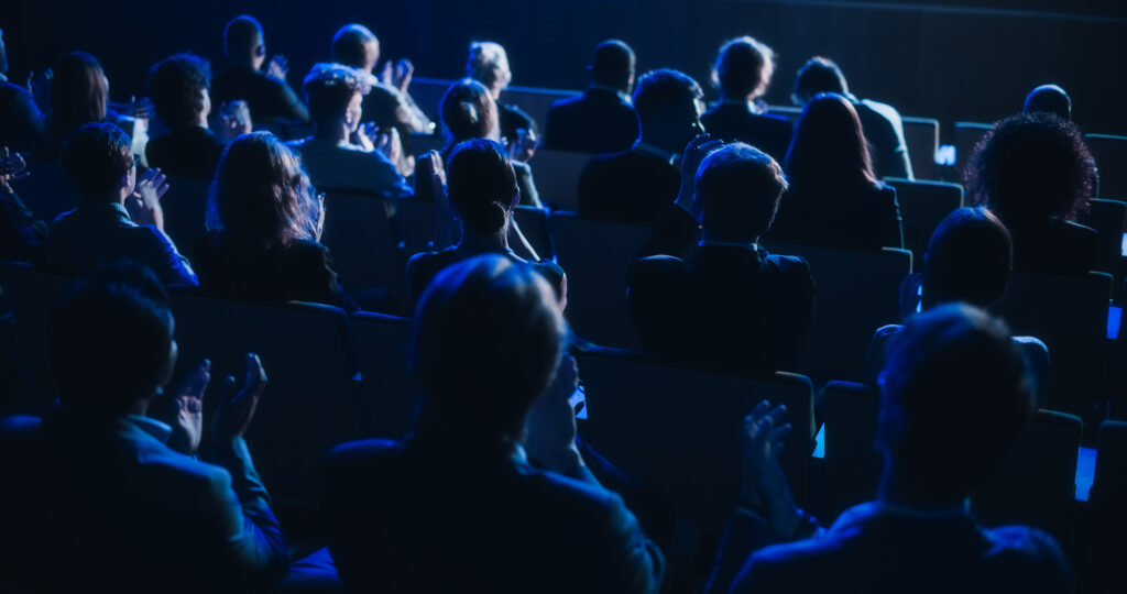 Yleisöä sivusta selin istumassa hämärässä auditoriossa.