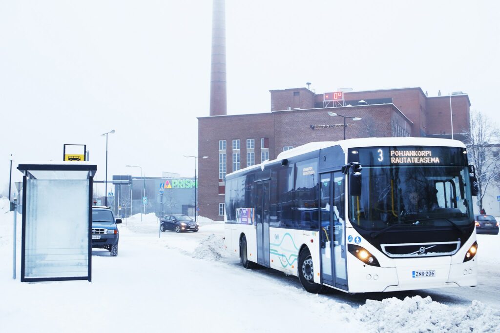 A bus stopped at a stop in a winter landscape with a power plant building in the background.