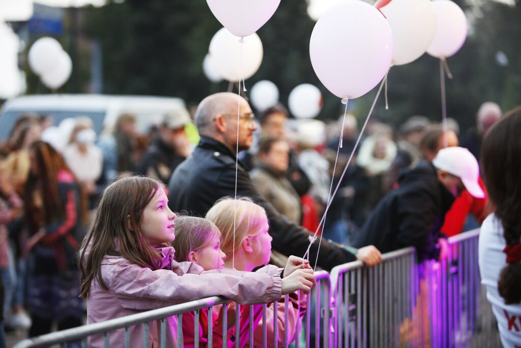 Väkijoukossa ulkotapahtumassa kolme lasta nojaa tapahtuman turva-aitaan eturivissä. Heillä on käsissään ilmapallot.