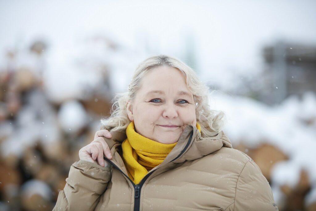 Anja Akkanen utanför klädd i en blek topprock med en snöig trähög i bakgrunden.