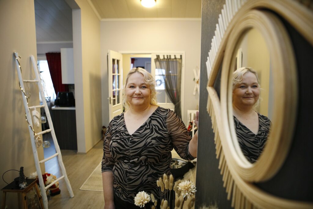 Anja Akkanen smiles, the hallways and kitchen of her home in the background. The mirror reflects Anja's image.