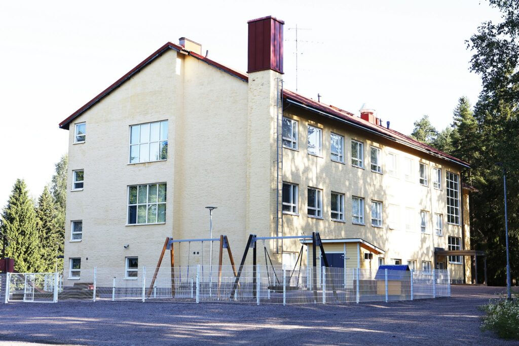 Yellow three-story school building, fenced yard with swings. Forest in the background.