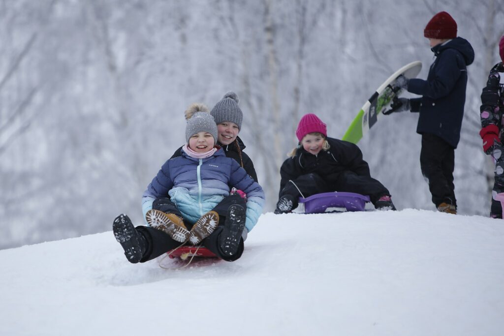 Kaksi pulkallista lapsia laskemassa mäkeä lumiselta kumpareelta, toisessa pulkassa isompi lapsi pitää pienempää sylissään. Mäen päällä seisoo kaksi lasta, joista toisella pehmoliukuri kädessään.