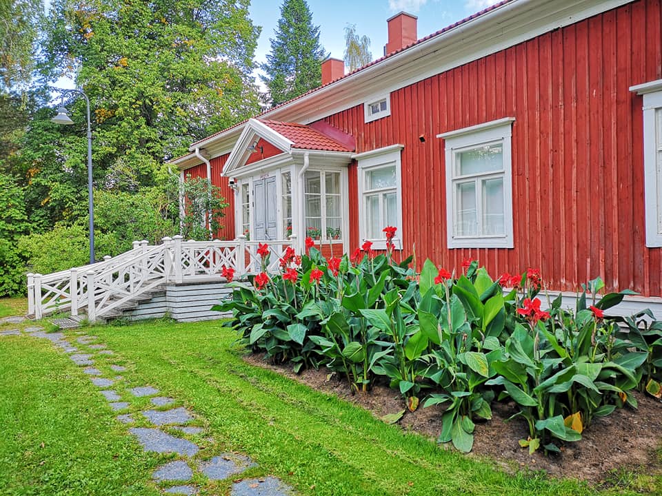 Vanhan punaisen puutalon julkisivu kesäisessä vehreydessä, kukkapenkki talon edustalla.