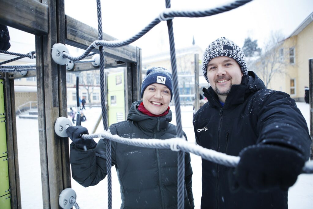 A woman and a man wearing winter coats and beanies are smiling by the rope lattice of the playground, both holding the rope with one hand.