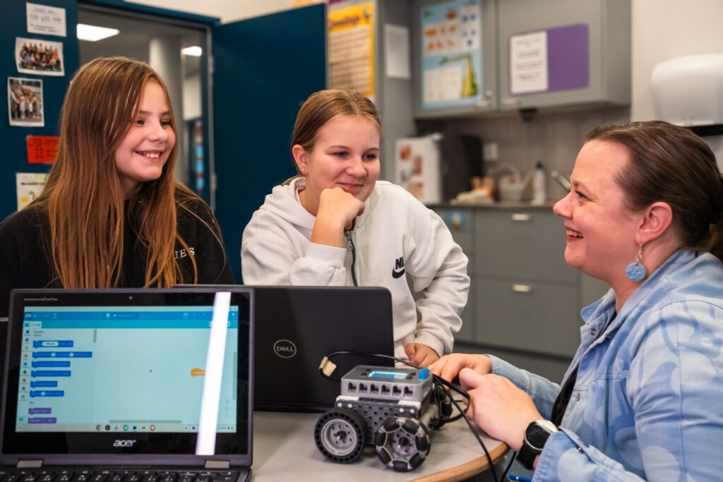 Två leende skolflickor och en lärare vid bordet. Bordet har två skärmar och en robot med hjul.