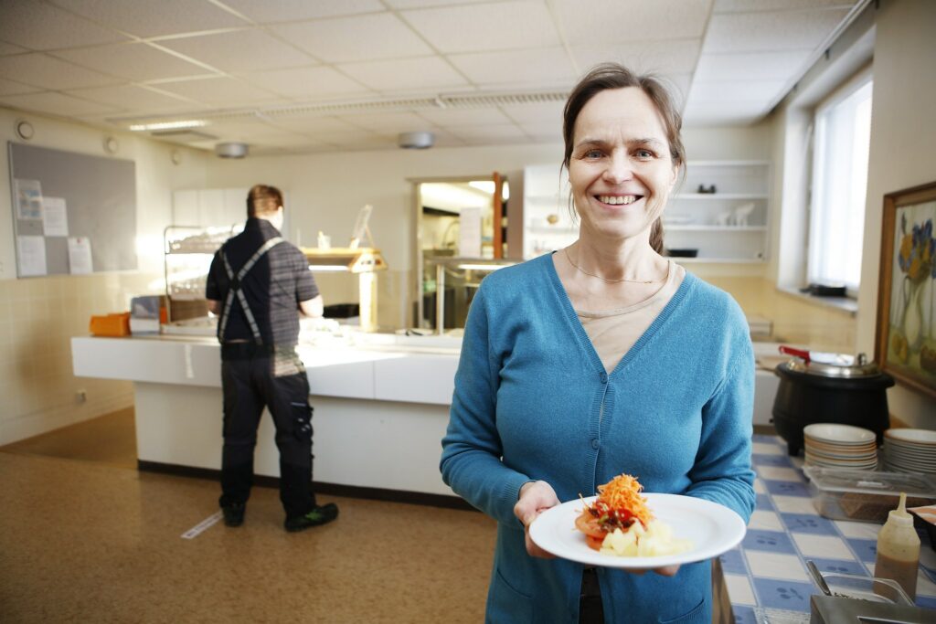In the foreground, a smiling woman with a plate in her hand. In the background, the restaurant's lunch line.