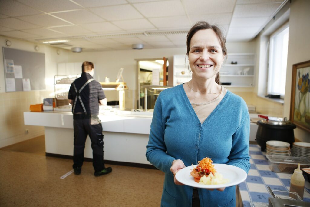 I förgrunden en leende kvinna med en tallrik i handen. I bakgrunden restaurangens lunchkö.