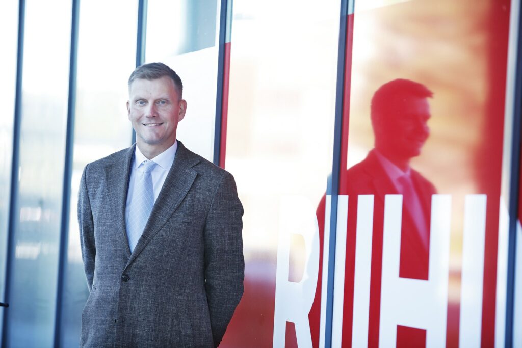 A man is standing in a suit outside in front of a glass wall, smiling. The glass wall has red tape and the visible text Riihi.