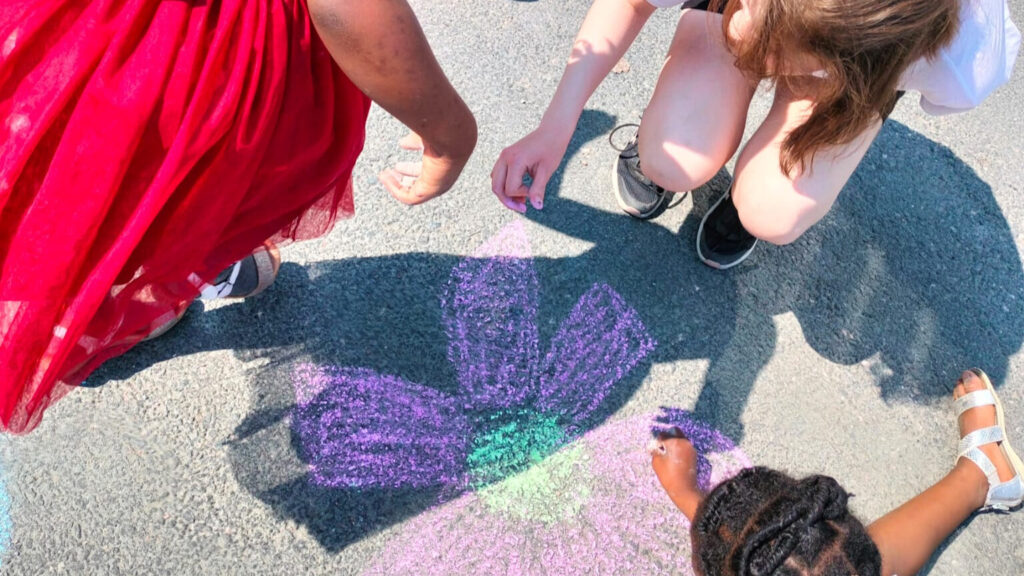 Asvaltilla väriliiduilla piirretty kukka ja kahden lapsen kädet piirtää liiduilla kukkaa. Lapsilla eri ihonvärit.
