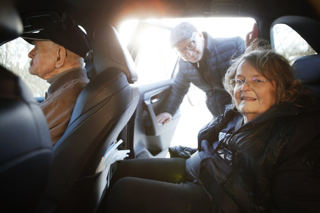 A man peeks into a car from the back door, where a smiling woman is sitting in the back seat and a man is in the front seat.