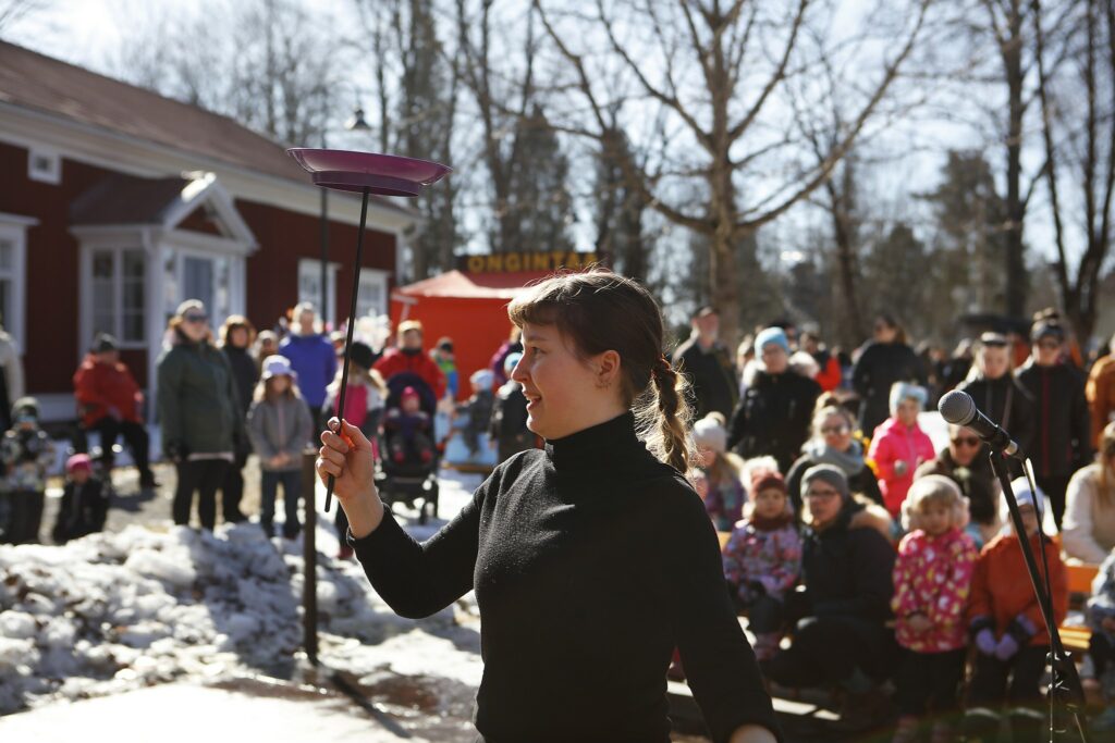 Paljon lapsia ulkona pihalla katsomassa esitystä, jossa nainen kävelee tikku kädessä, jonka nokassa on lautanen. Vieressä mikrofoni. Taustalla punainen puutalo.