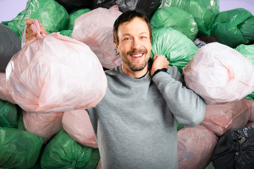Hymyilevä mies makaamassa selällään värikkäiden roskapussien keskellä, yksi pussi kädessä nostettuna ylös ilmaan.