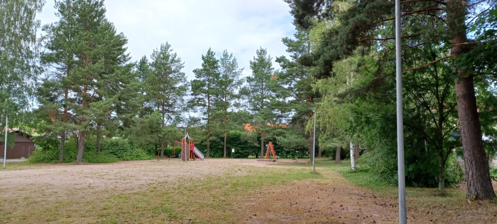 A sand square bordered by trees, with a sandbox, swings and a slide at the back.