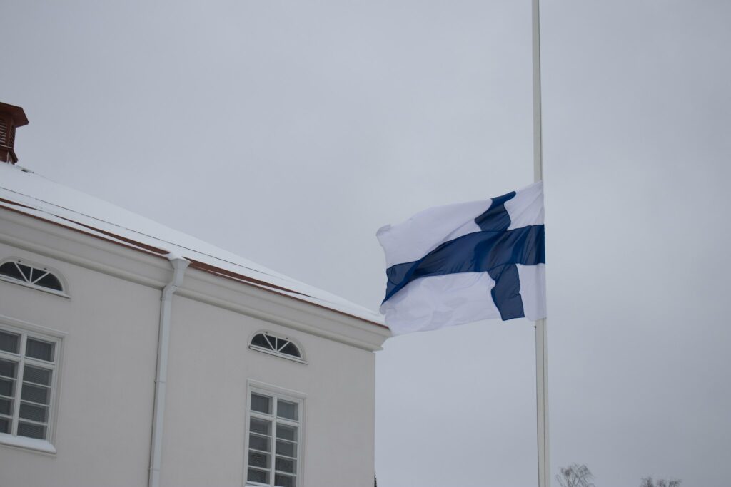 Suomen lippu liehumassa puolisalossa harmaata taivasta vasten. Vaalea rakennus osin taustalla.