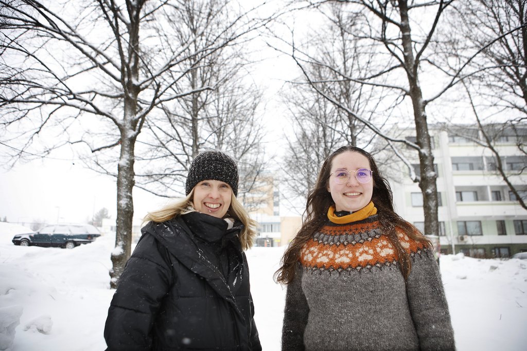 Två kvinnor ler glatt bredvid varandra i ett snölandskap med björkar och ett hyreshus i bakgrunden.