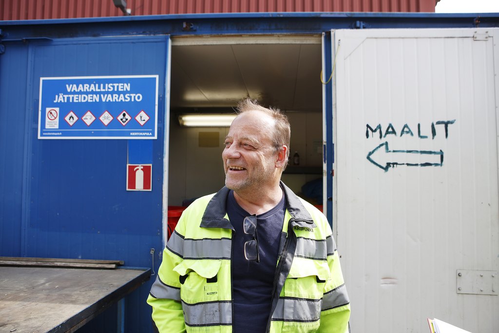 En man tittar till vänster med ett godmodigt leende framför en blå container i sin iögonfallande jacka. Dörren till containern är öppen och en pil och texten Maalit är ritade med en markör på luckan.