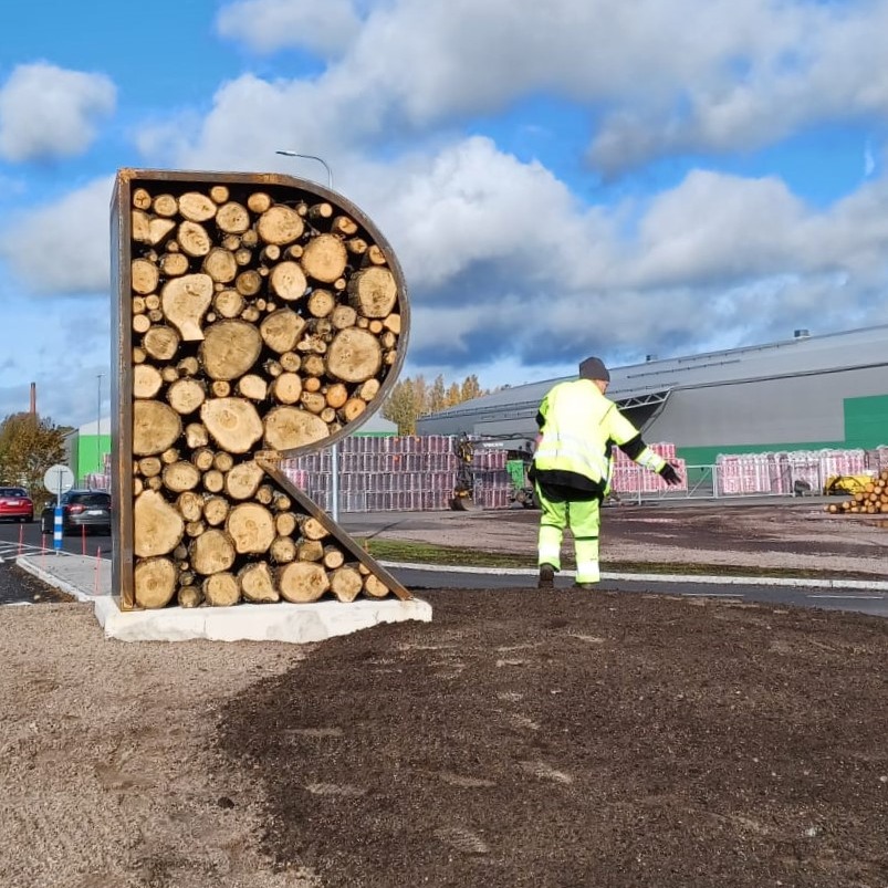 A large "bug hotel" in the shape of the letter R, where the metal frames of the letter R are filled with halos. Around Ötökkähotell there is dirt on the ground, one street worker in attention clothes on his back and a light blue sky with gray-white clouds.