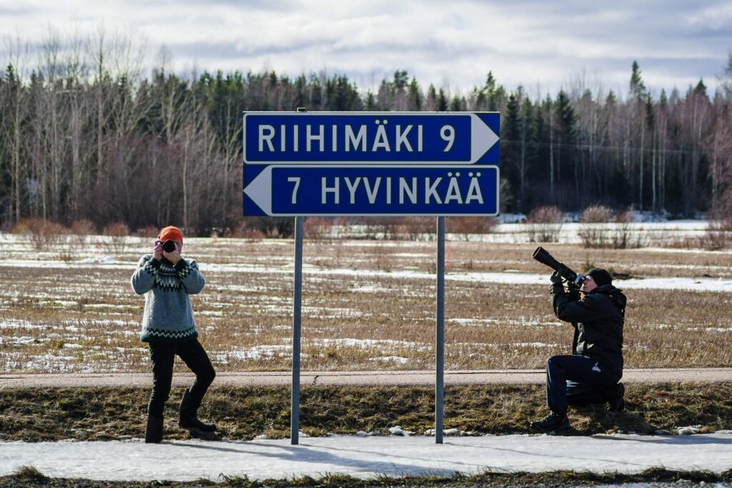 Keväisen pellon laidassa tienviitat Riihimäelle sekä Hyvinkäälle. Kaksi henkilöä poseeraa tienviittojen vieressä