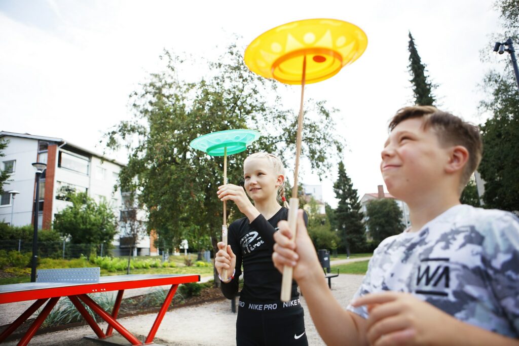Kaksi lasta pyörittää sirkustemppuvälineitä ilmassa kesäisessä puistossa.