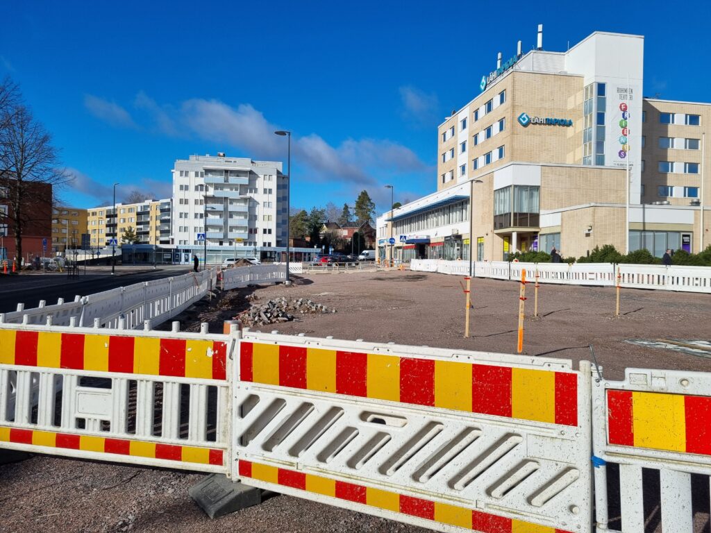 Construction site fence, behind which is a sandy square. In the background, light apartment buildings against the blue sky.