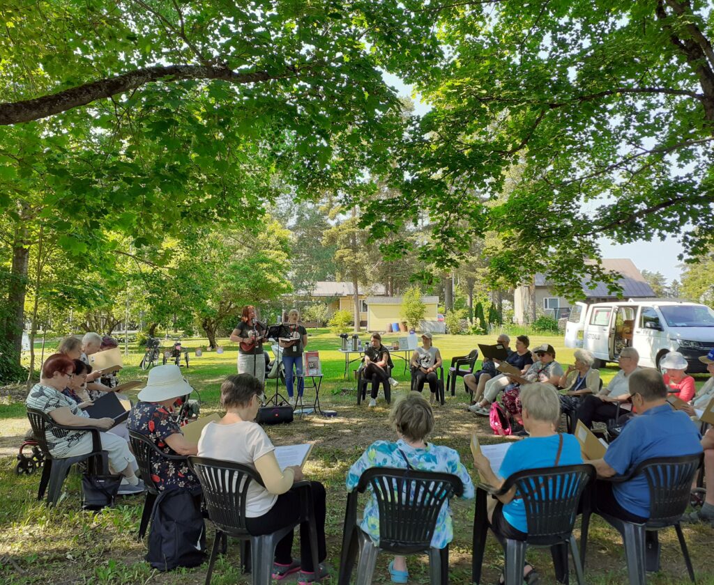 I en grön park på gräset sitter människor i en stor cirkel på plaststolar med öppna pärmar i händerna. Två personer som spelar instrument i ringen.