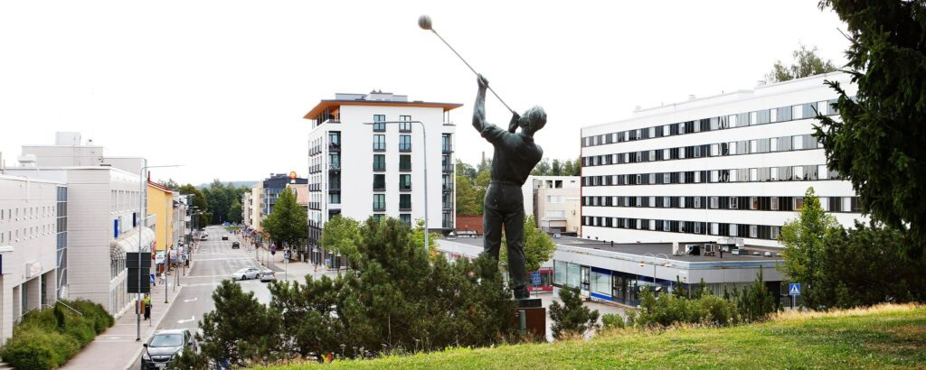 En staty av en glasblåsare på en kulle vid foten av en barrbuske, med centrala hyreshus i bakgrunden.