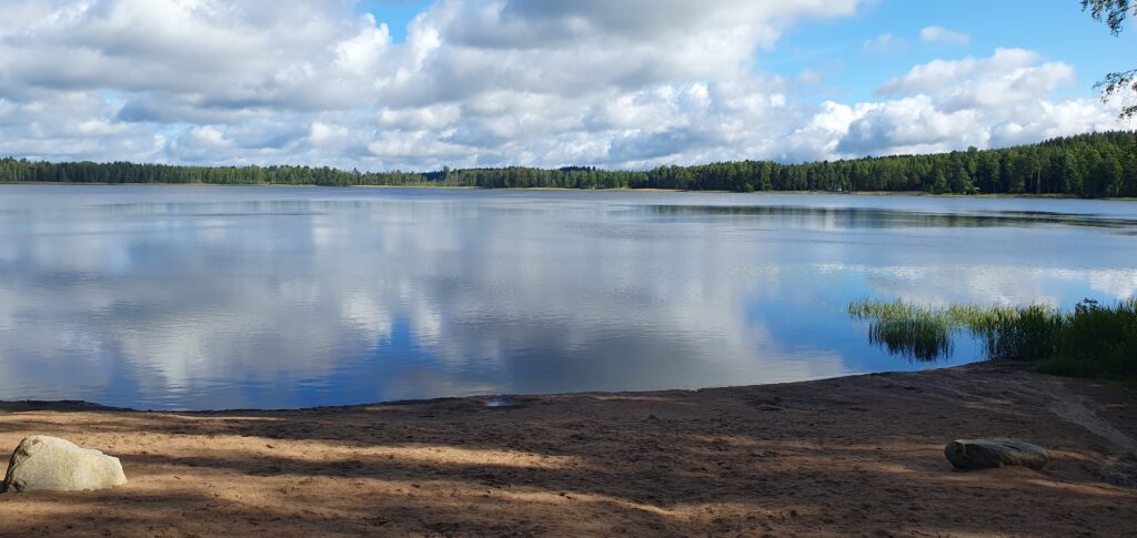 Hiekkaranta ja järvi, josta heijastuu pilvet. Järveä reunustaa metsä.