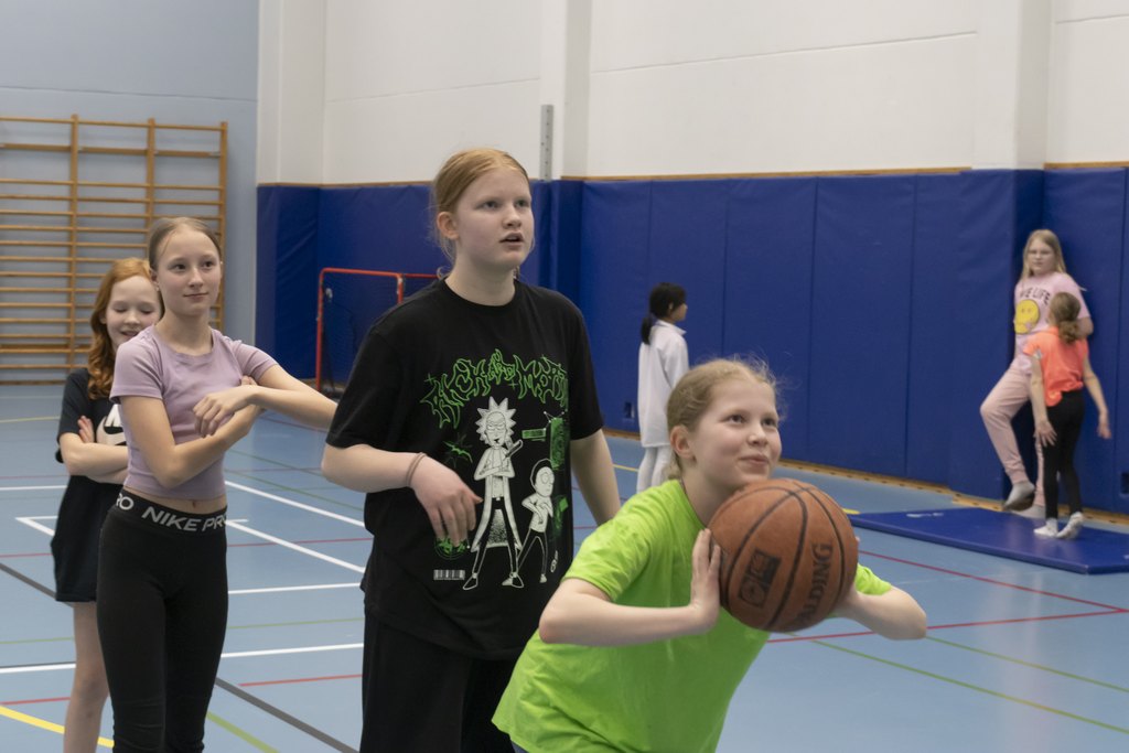 Neljä tyttöä jonossa, joista ensimmäinen heittämässä palloa koriin liikuntasalissa.