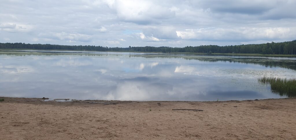 An empty beach, fine beach sand and clouds are reflected in the water of the lake. The lake is bordered by a forest.