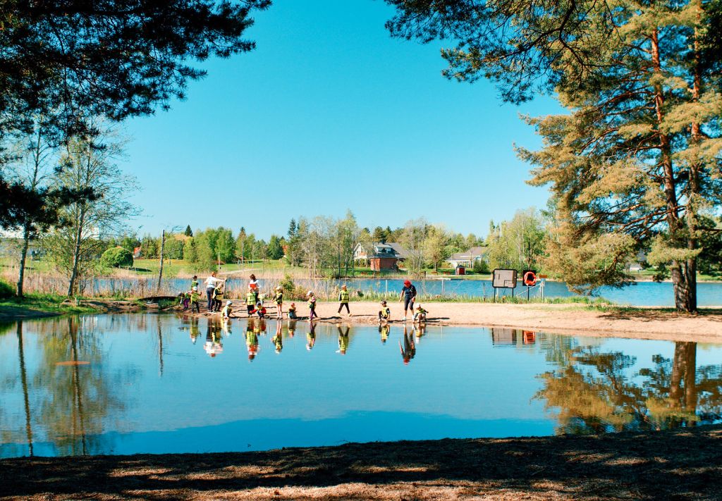A lagoon-like lake, with a group of children in attention vests with adults on the far shore. The picture is framed by a couple of large pine trees. Pale blue sky, calm lake surface.