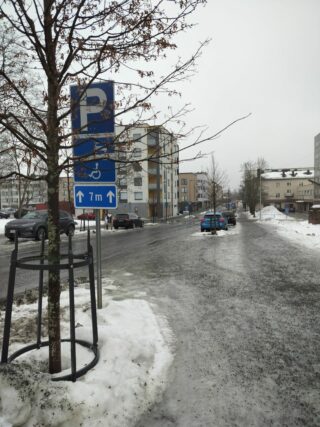 Luminen ja jäinen katunäkymä kirjaston edessä olevasta tiestä: pysäköintimerkki kertoo inva-paikan sijainnista. 
