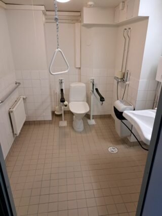 Vessanpöntön vieressä on hälytin ja tukikaiteita, vessassa myös roskakori ja käsienpesuallas.