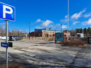 Pysäköintimerkki parkkipaikan vieressä, taustalla iso rakennus.