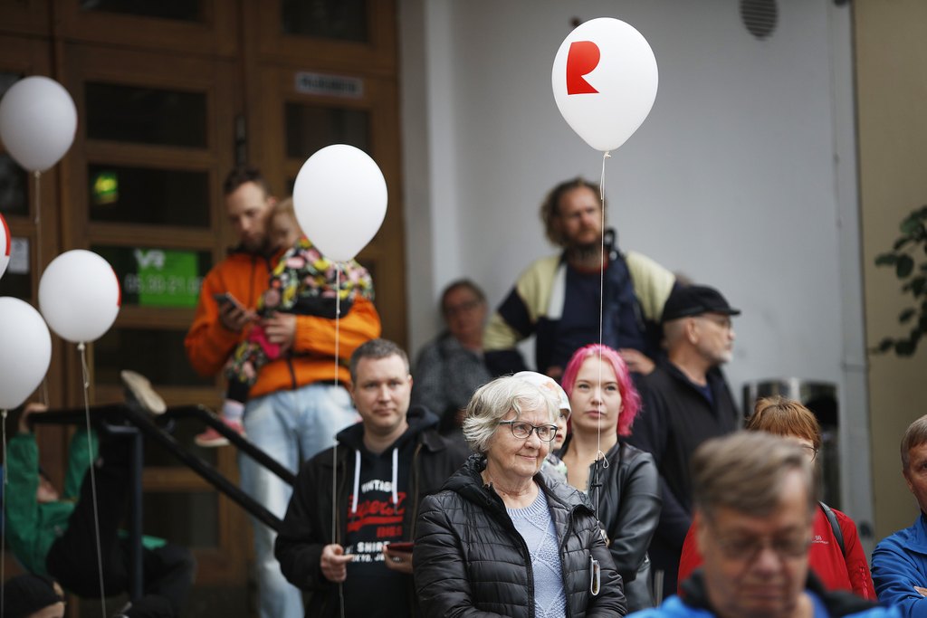 Eri ikäisiä ihmisiä kokoontuneena rakennuksen edustalle. Ilmassa kaksi ilmapalloa, joissa punainen R-kirjain.