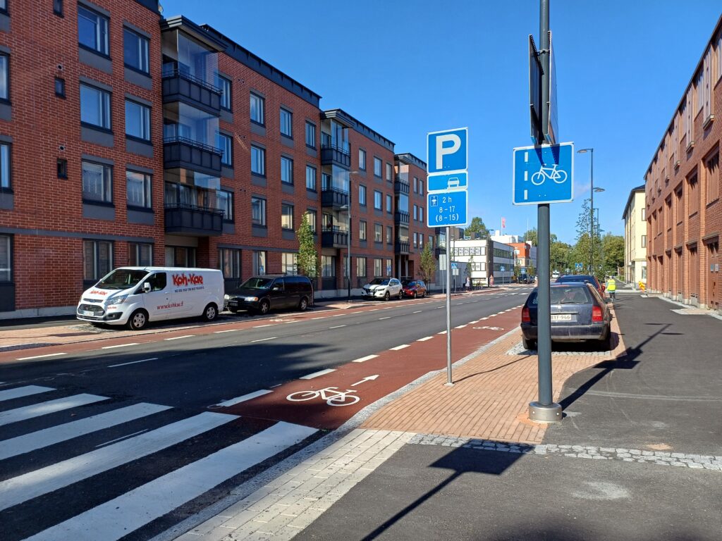 Autotie keskustassa, etualalla suojatie, pyöräkaistaa ja kadunvarsipysäköintipaikat. Katua reunustaa muutaman kerroksen korkeat kerrostalot. Pilvetön kirkas taivas.