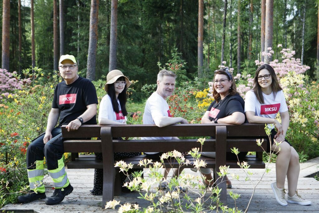 Viisi naista ja miestä istumassa puiston penkillä hymyillen t-paidoissaan kesäpäivänä. Taustalla metsää ja kukkia.