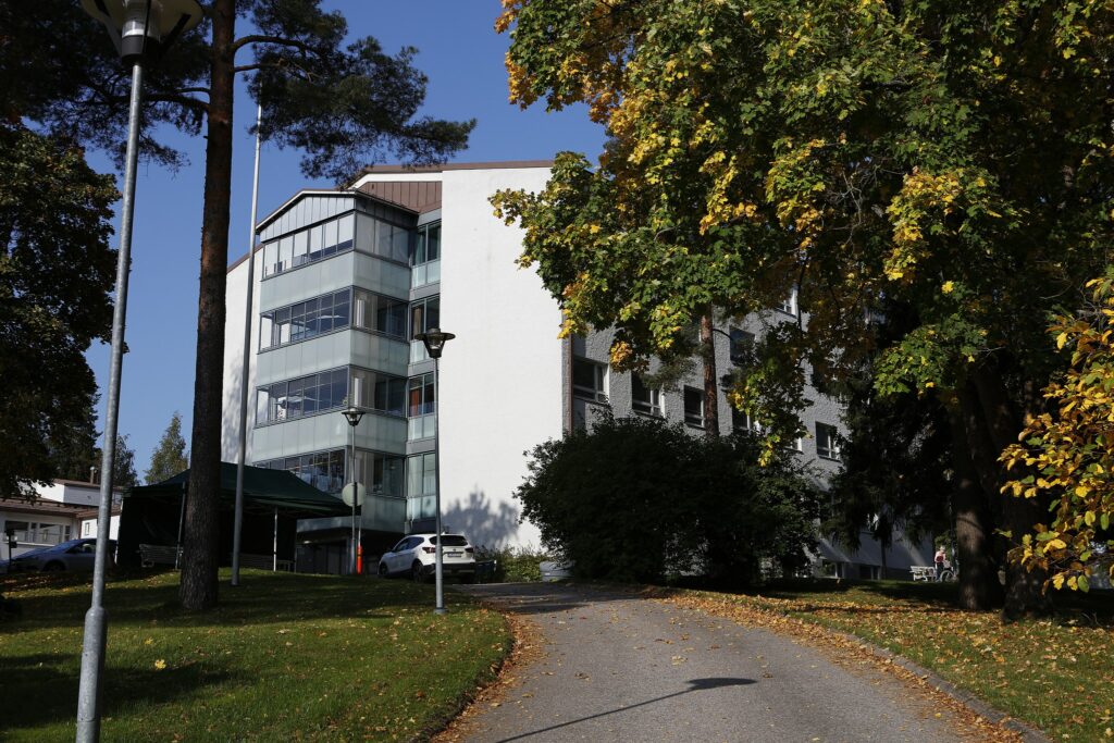 Valkoinen uudehkon näköinen monikerroksinen talo vehreässä puistomaisemassa. Talolle johtaa asfalttitie.
