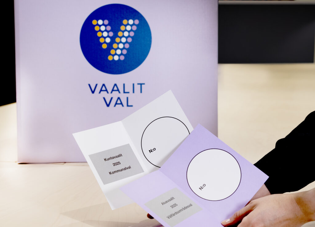 Vaaliuurna pöydällä sekä ihmisen käsissä kaksi äänestyslipuketta, joista toinen on valkoinen ja toinen on violetti.