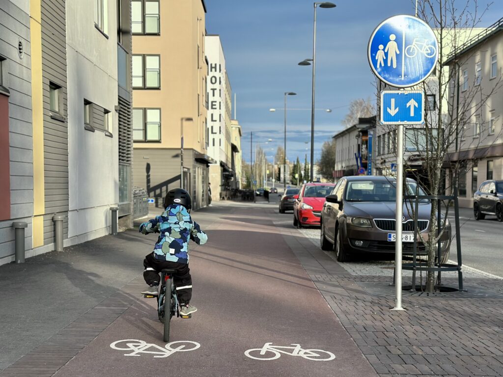 Pieni lapsi pyöräilemässä kamerasta poispäin keskustan kevyen liikenteen väylällä. Kerrostaloja molemmin puolin kevyen liikenteen väylää ja autotietä.