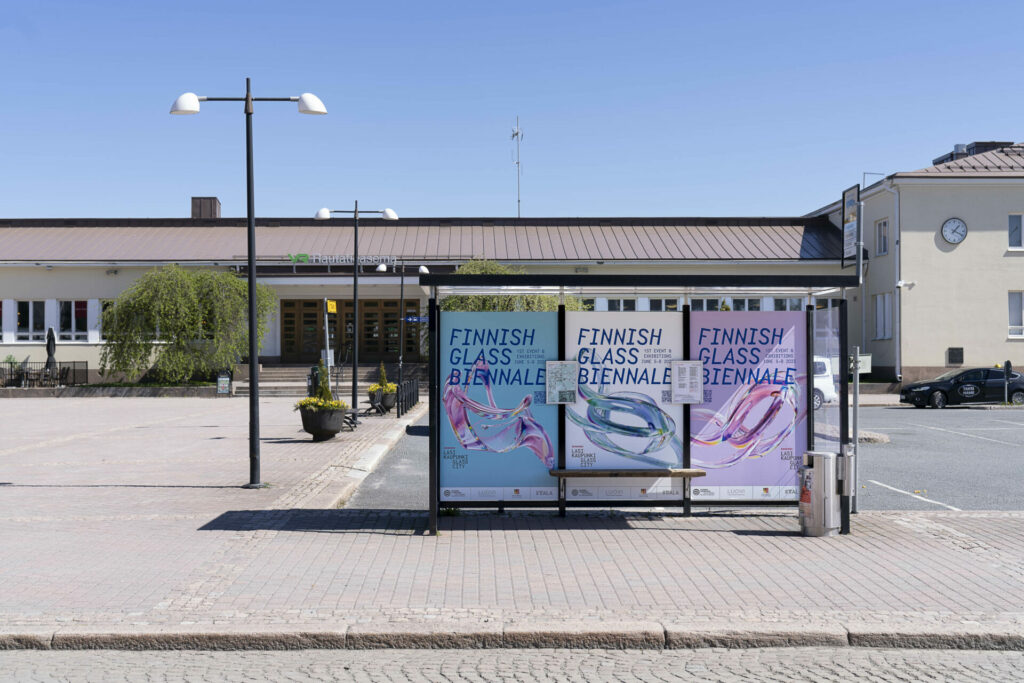 Rautatieasemarakennuksen ja aukion edessä bussikatos, jonka seinillä värikkäät mainokset. joissa lukee Finnish Glass Biennale.